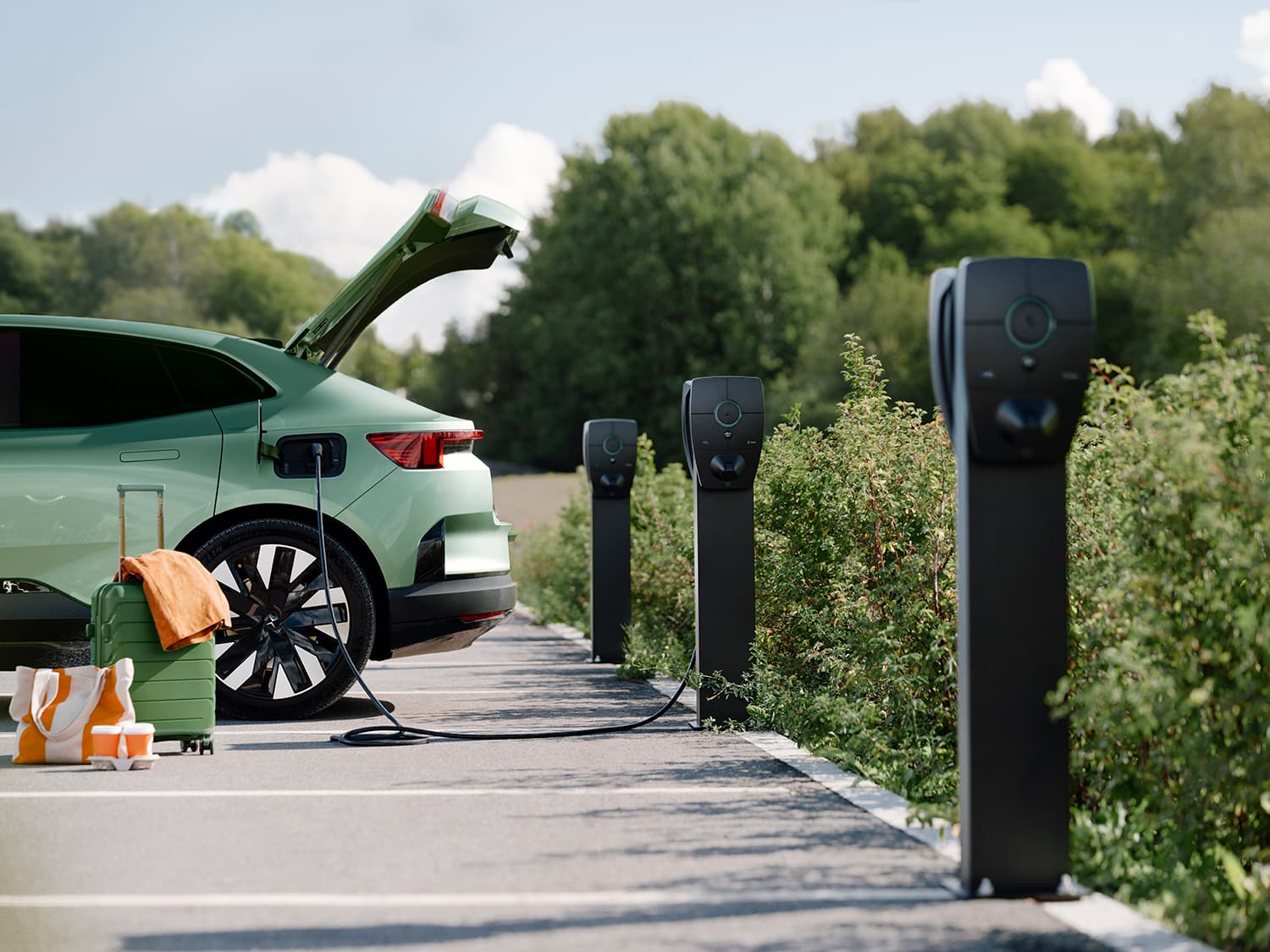 Grünes Auto lädt an Zaptec Pro Ladestation auf Säule, drei Ladestationen sichtbar, Koffer und Tasche am Boden, Kofferraum offen.