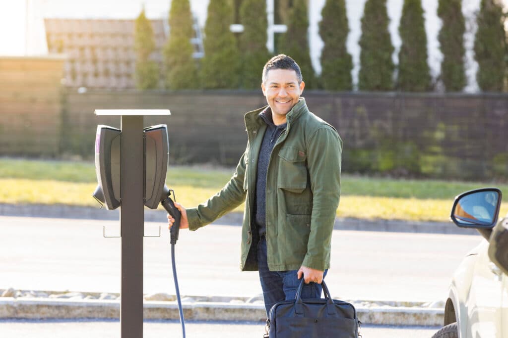 En smilende mann i grønn jakke lader elbilen sin med en Zaptec Pro-lader utendørs. Han holder en blå bag i den ene hånden og ladekabelen i den andre, med solen som lyser opp scenen.