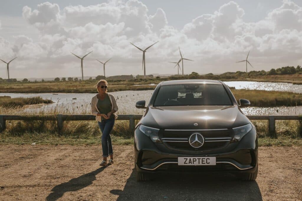 En kvinne går bort fra en svart Mercedes elbil med registreringsskiltet "ZAPTEC", parkert ved et våtmarksområde. I bakgrunnen sees flere vindmøller og et åpent landskap under en dramatisk skyhimmel.