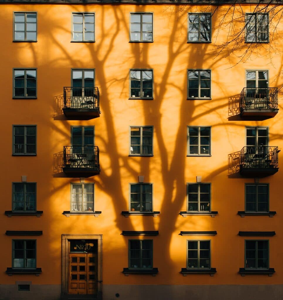 En gul fasade på en bygård bader i sollys, med skyggene av tregrener dramatisk projisert over bygningen. Flere balkonger og vinduer vises i et symmetrisk mønster.