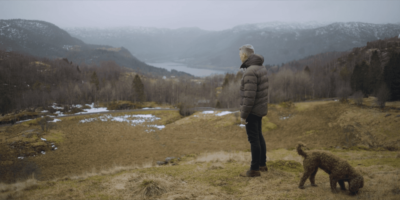 en man i mörka kläder står tillsammans med en brun hund i ett bergigt landskap med gröna områden och små snöfläckar
