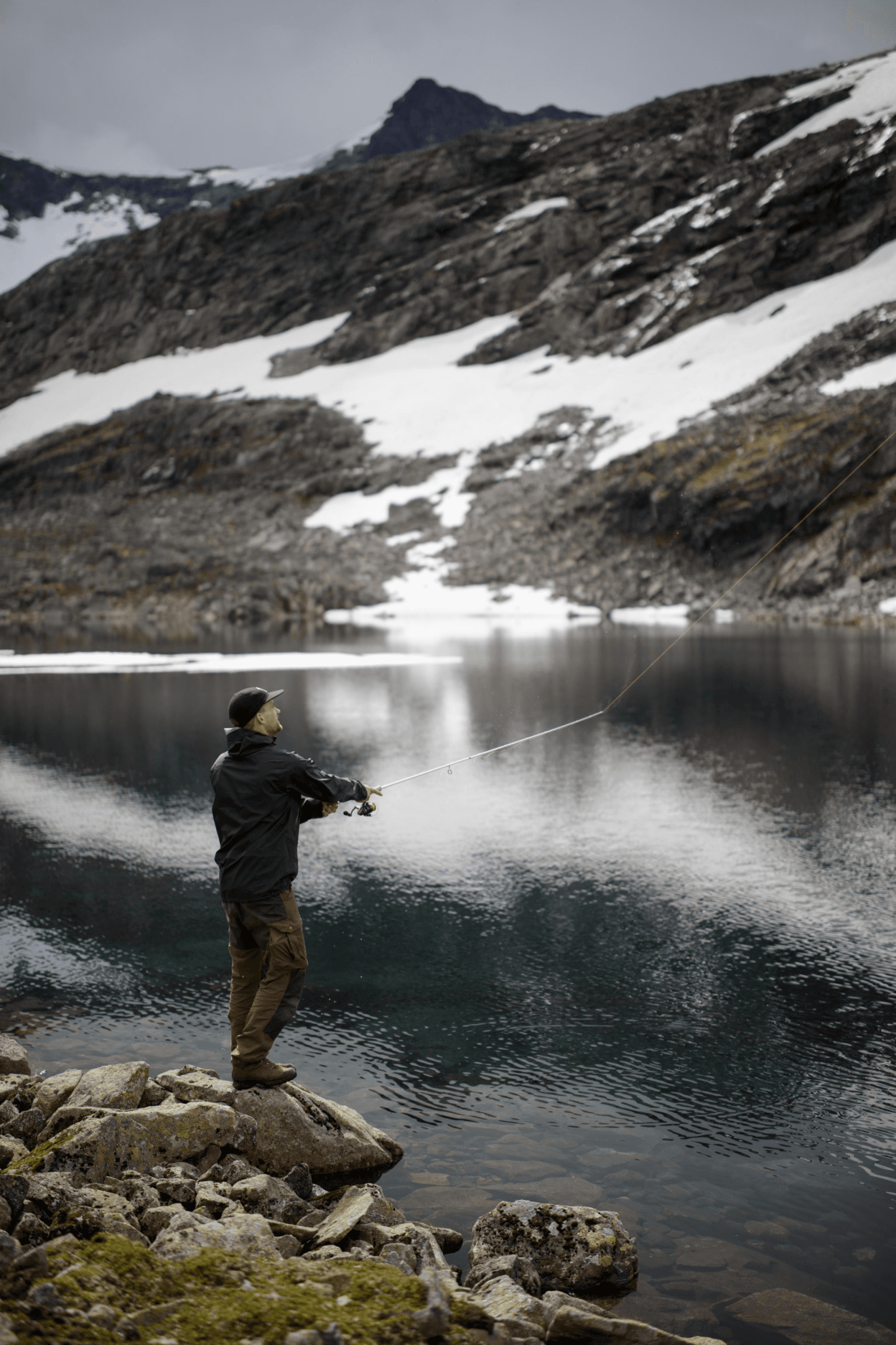 En man med mörka kläder står på en sten vid kanten av ett spegelblankt vatten och kastar ut en fiskelina i vattnet. I bakgrunden tonas ett fjäll upp som är delvis täckt av snö, fjället reflekteras ned i vattnet och lämmanar ett grå vitt mönster på vettenytan.