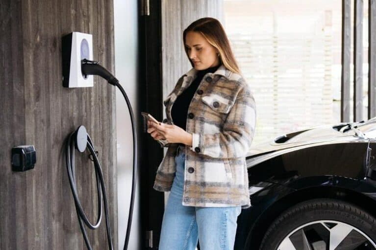 en kvinna med mörkthår, jeans och beige-vit overshirt står med vin telefon vid en vit Zaptec Go laddare som är installerad på ett modernt hus i mörkt trä.