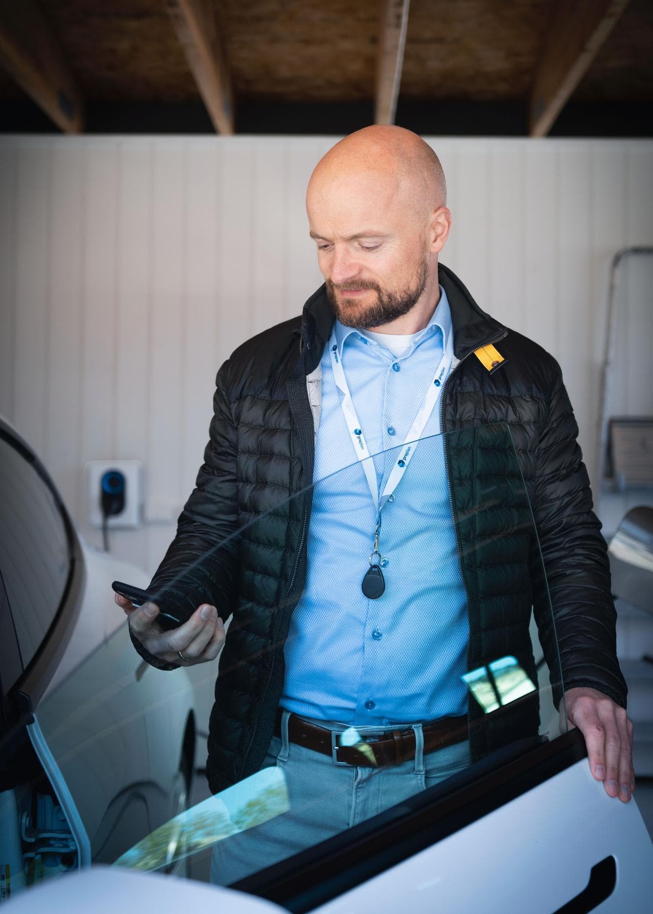 En mann står ved en hvit elbil i en garasje og ser på mobiltelefonen sin. I bakgrunnen vises en Cloud White Zaptec Go-lader montert på veggen.