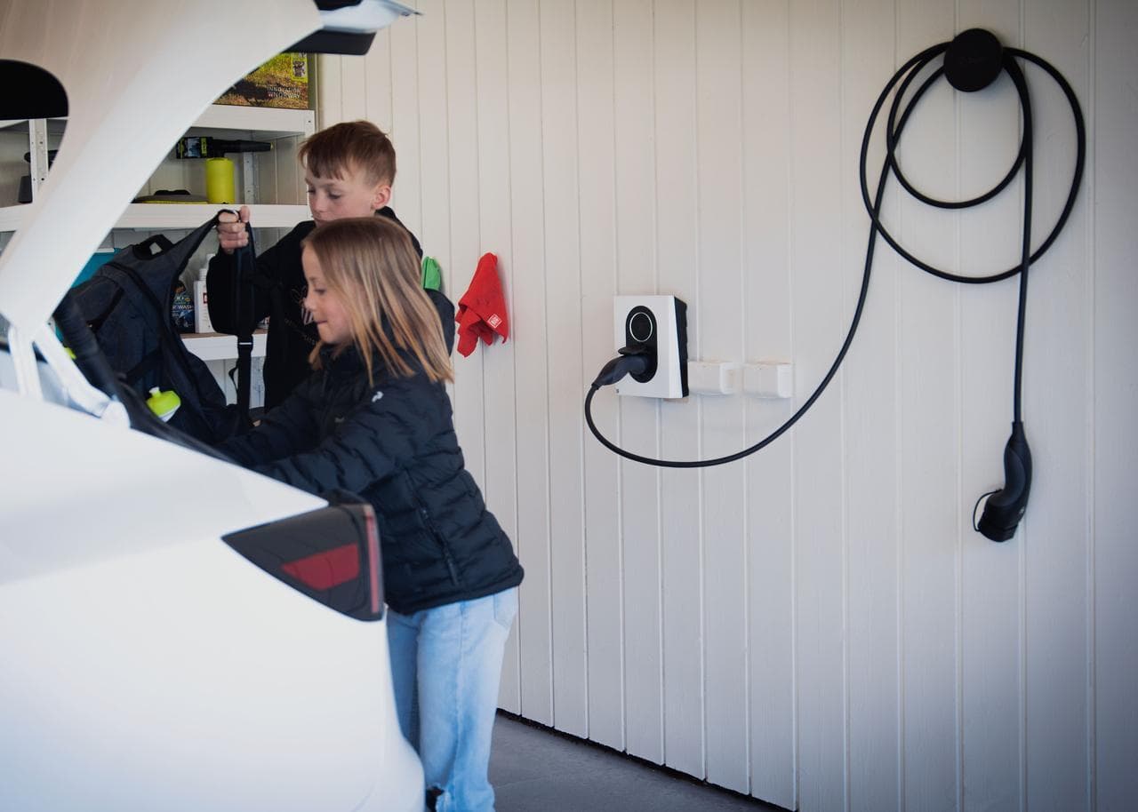 två barn packar in väskor i bagageluckan på en vit elbil som står i ett vitt garage, på väggen sitter en vit Zaptec Go laddbox med en laddkabel inkopplad i laddaren som hänger prydligt ihoprullad på kabelhållaren Zaptec Chill