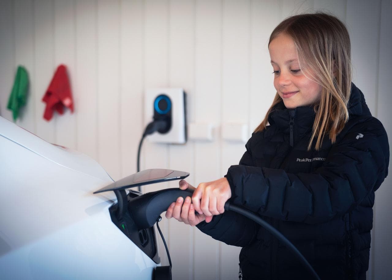 En smilende jente i svart jakke lader en hvit elbil i en garasje. I bakgrunnen vises en Cloud White Zaptec Go-lader montert på en hvit trevegg.