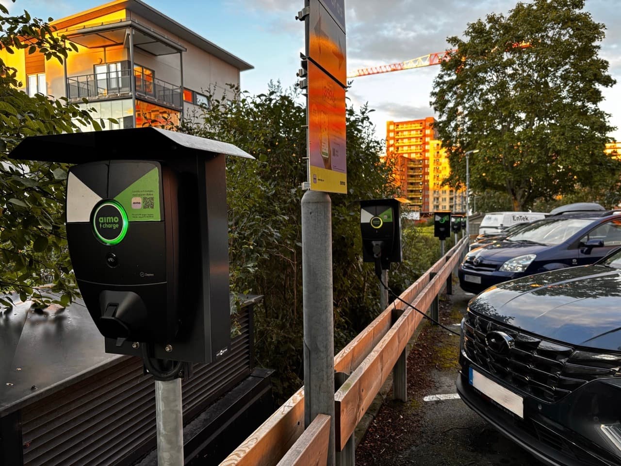 En rad Zaptec Pro-laddare är monterade på stolpar längs en bostadsgata där flera bilar står parkerade. Det varma ljuset från solnedgången lyser upp husen och bilarna och framhäver områdets moderna och hållbara infrastruktur.