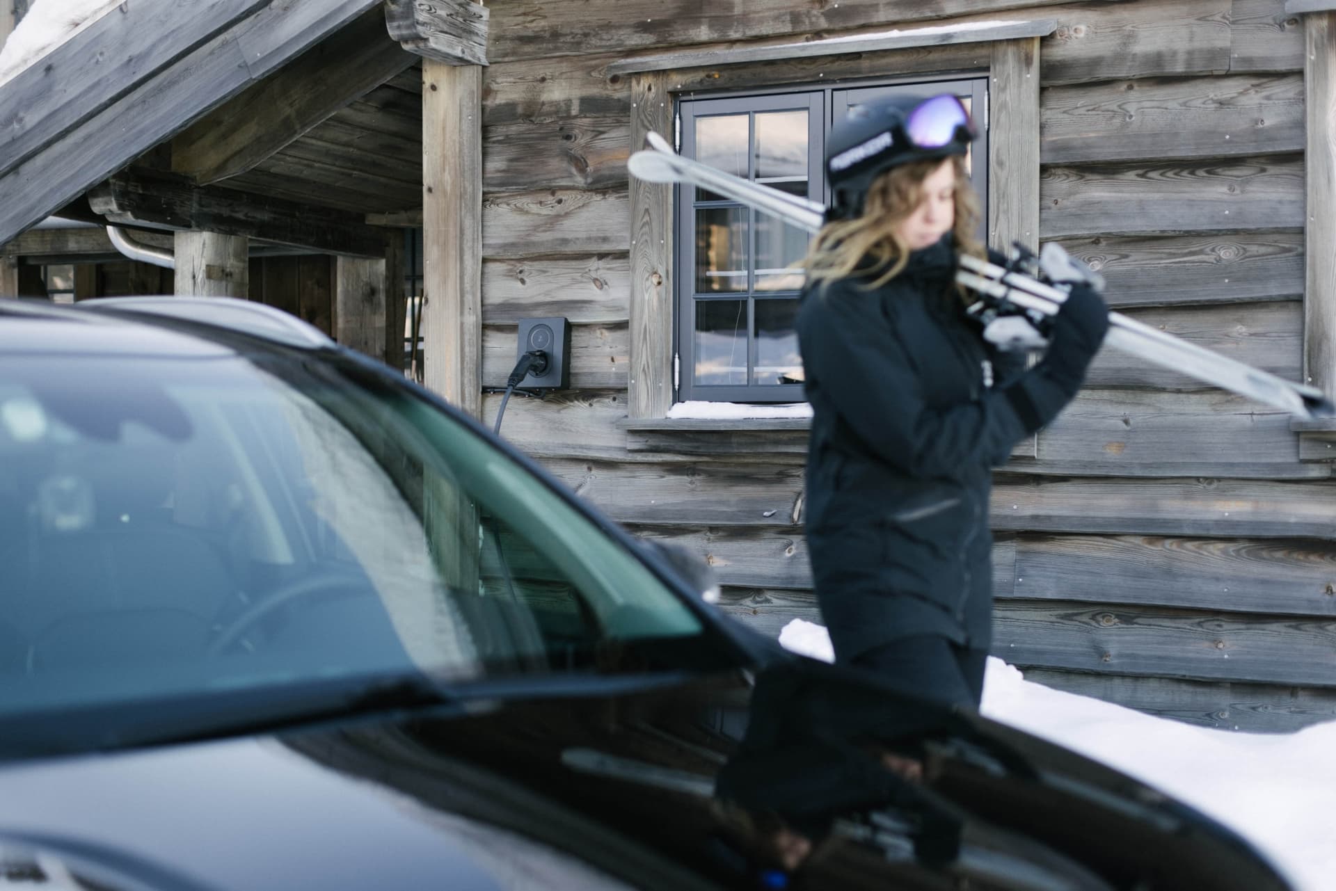 Kvinna som bär på skidor vid en timmerstuga. Framför henne står en svart bil som laddar med en Asphalt Black Zaptec Go laddare som sitter på väggen på det timrade huset.