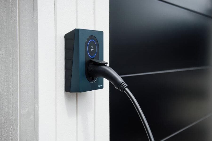 En Midnight Blue Zaptec Go-lader er montert på en hvit yttervegg og lader en elbil. Veggen ved siden av er sort med horisontale linjer, og ladekabelen er koblet til.