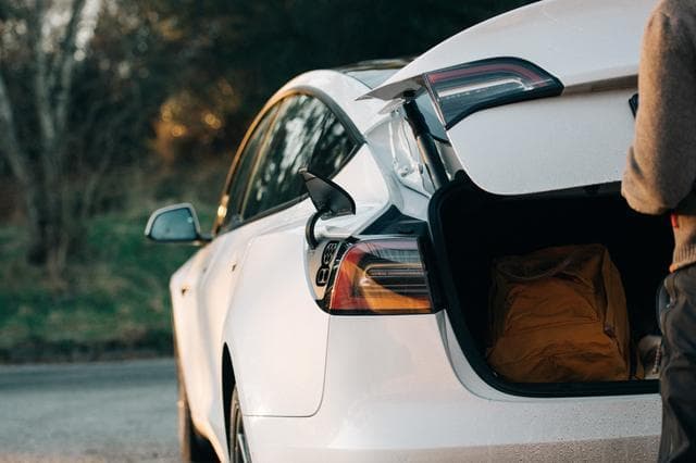 En vit elbil står parkerad, sidan en person i mörka kläder syns stänga bagageluckan, Det ligger en orange ryggsäck i bagageluckan och luckan för laddutag är öppet på bilen.