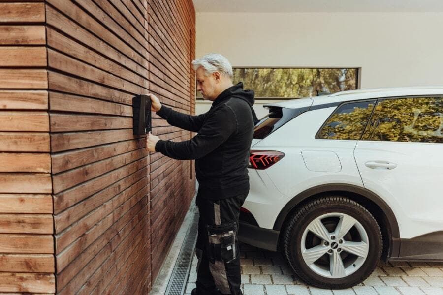 En mann i svart hettegenser installerer en Zaptec Go lader på et hus med vertikale trepaneler. En hvit elbil står parkert ved siden av på en steinlagt innkjørsel.