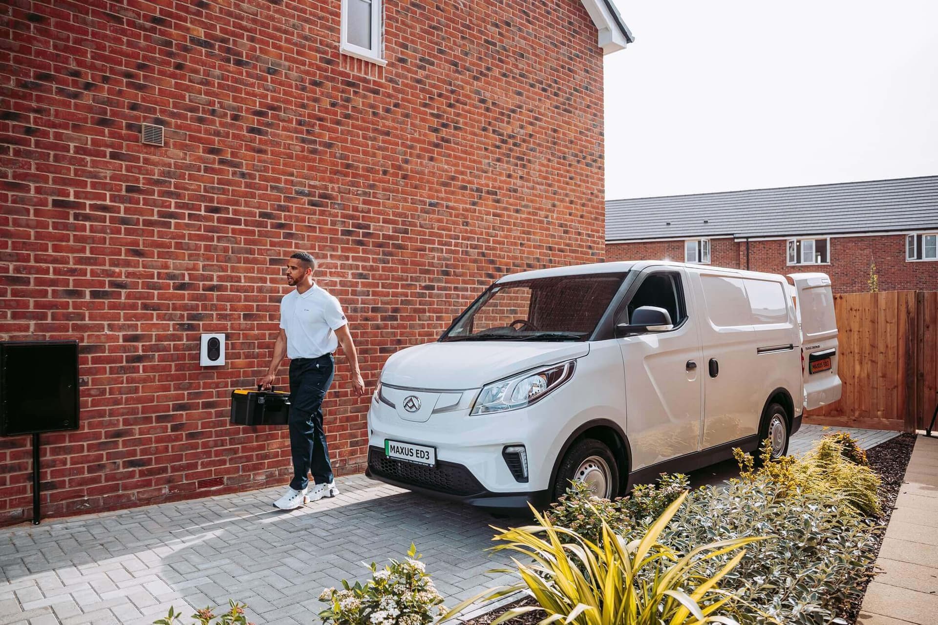 Une personne se tient devant un mur en briques à côté d’une borne de recharge murale noire, tandis qu’un utilitaire électrique blanc est garé dans l’allée.