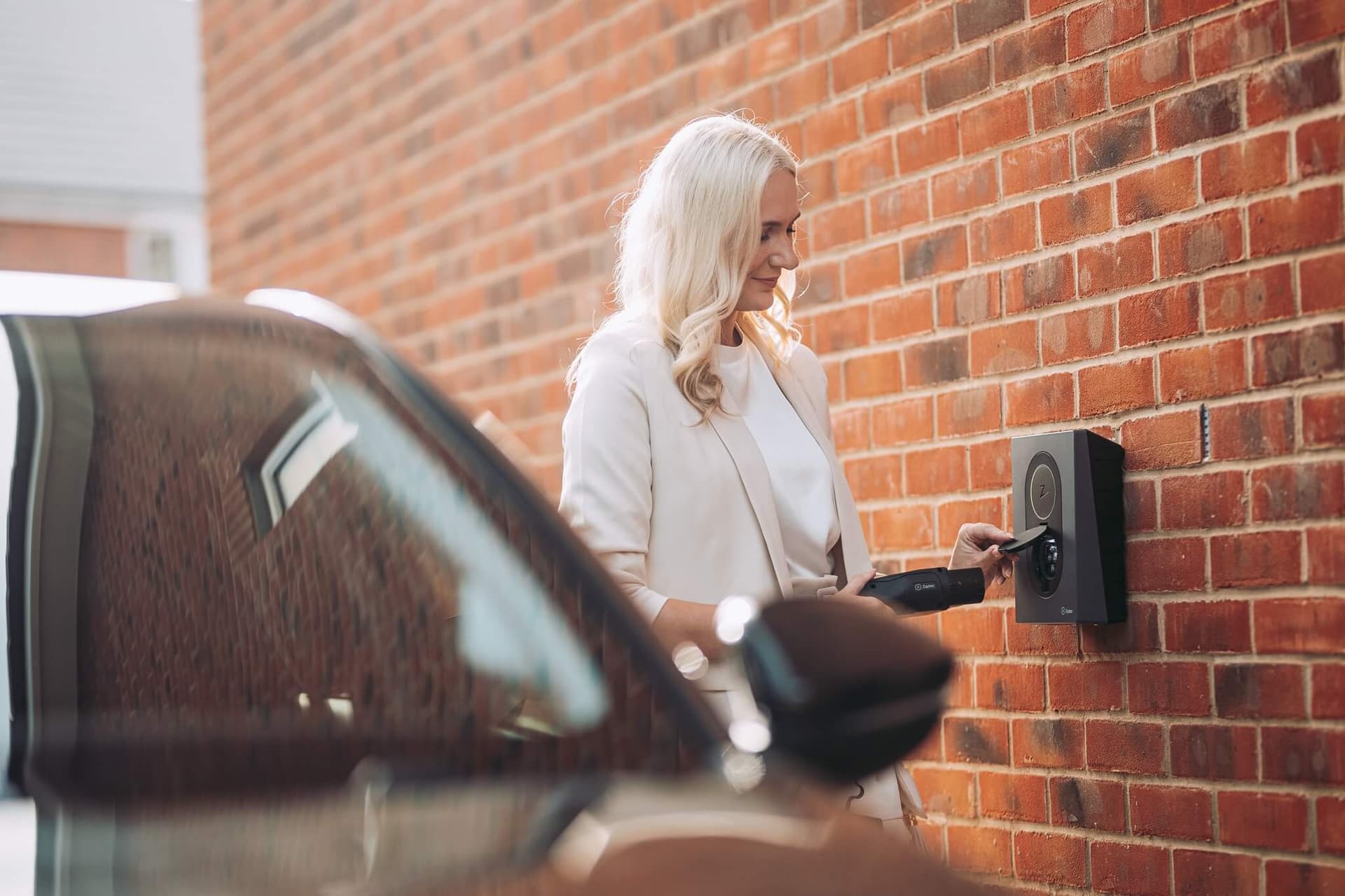 Une femme se tient près d’une borne de recharge murale, avec une voiture au premier plan.