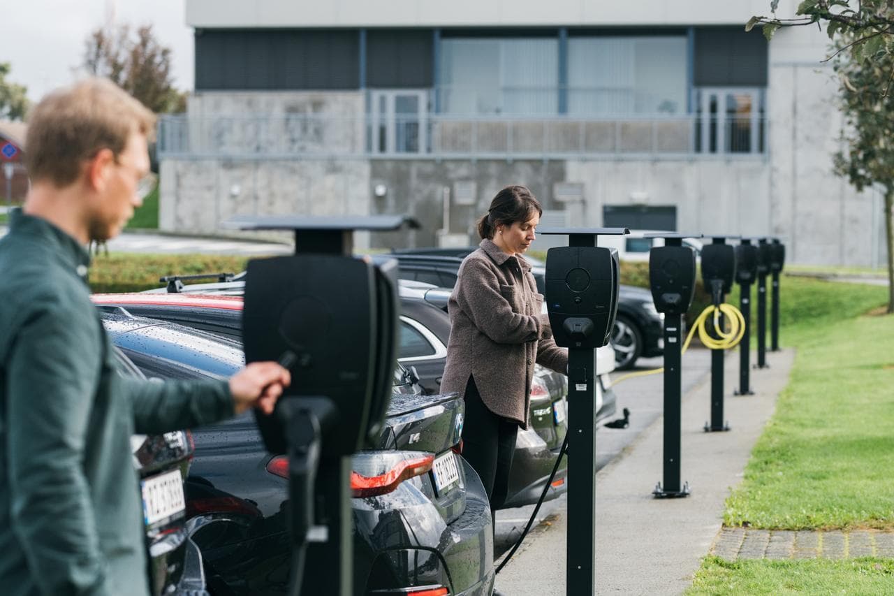 En parkeringsplats med Zaptec Pro MID laddare installerade på laddstolpar, flera bilar står parkerade och laddar. En kvinna och en man klädd i mörka färger står vis två av elbilsladdarna och startar laddning.