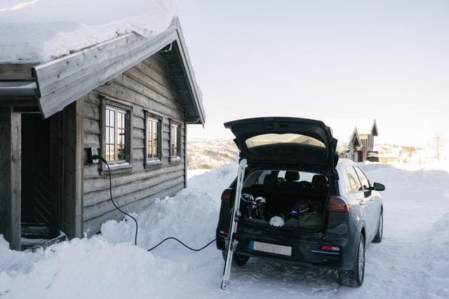 En svart elbil lades med en Zaptec Go-lader montert på veggen til en snødekt tømmerhytte. Bilens bakluke er åpen og et par ski stikker ut.