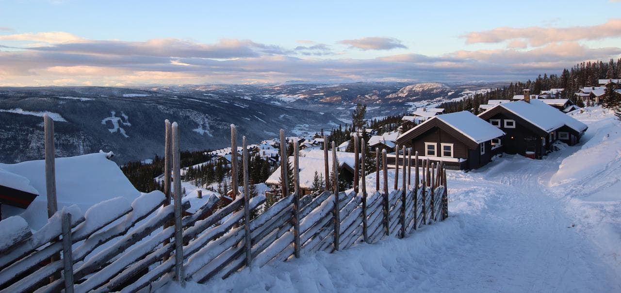 ett vinterlandskap med en gärdsgård och små bruna stugor på ett berg inbäddade i snö, man ser långt över snö och trätäckta fjäll med blå himmel och fluffiga moln i horisonten