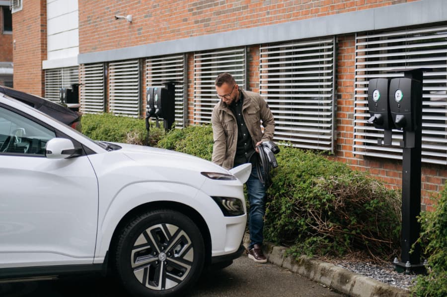 En man i en grön jacka och glasögon håller på att sätta i en Zaptec Pro laddare i framdelen på sin vita elbil som står parkerad vid en offentlig laddningsstation utanför en byggnad med tegelfasad.