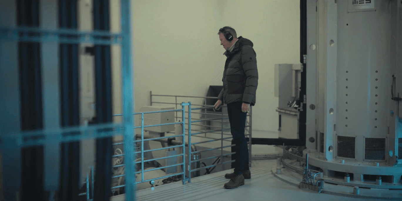 en man med mörka kläder och hörselkåpor står inne på ett kraftvärk och håller i ett stängsel