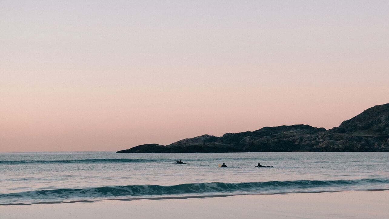 Tre surfere ligger på brettene sine i havet ved en rolig strand i solnedgang, med pastellfarget himmel og lave bølger. I bakgrunnen sees en lav klippeformasjon.