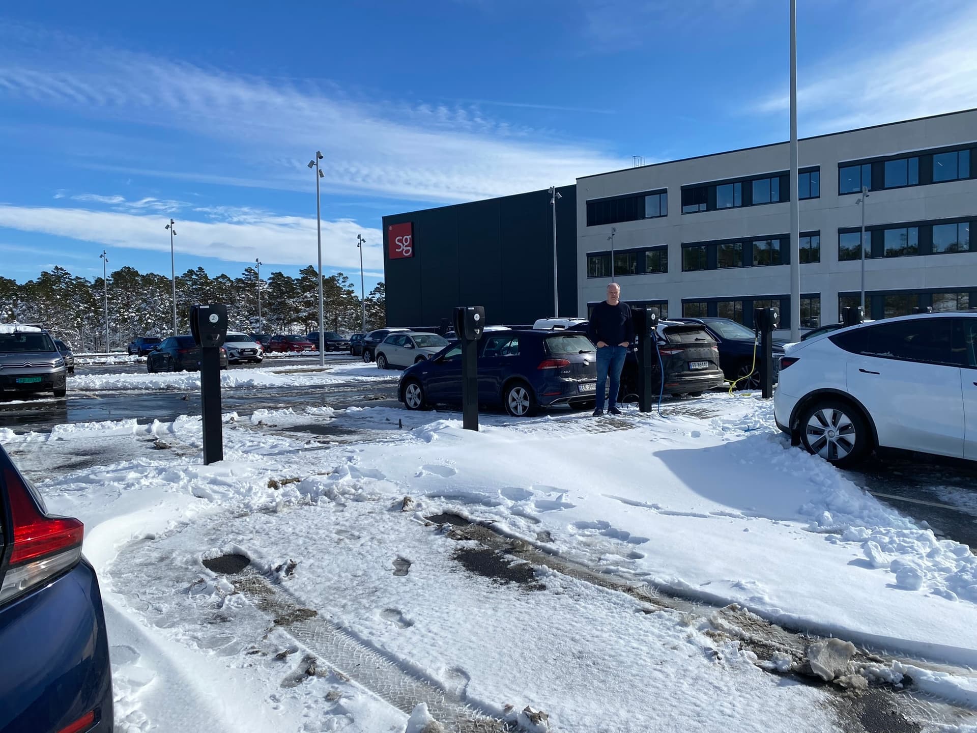 Flere elbiler lader med Zaptec Pro-ladere på en snødekt parkeringsplass utenfor en kontorbygning. Solen skinner fra en klar blå himmel.