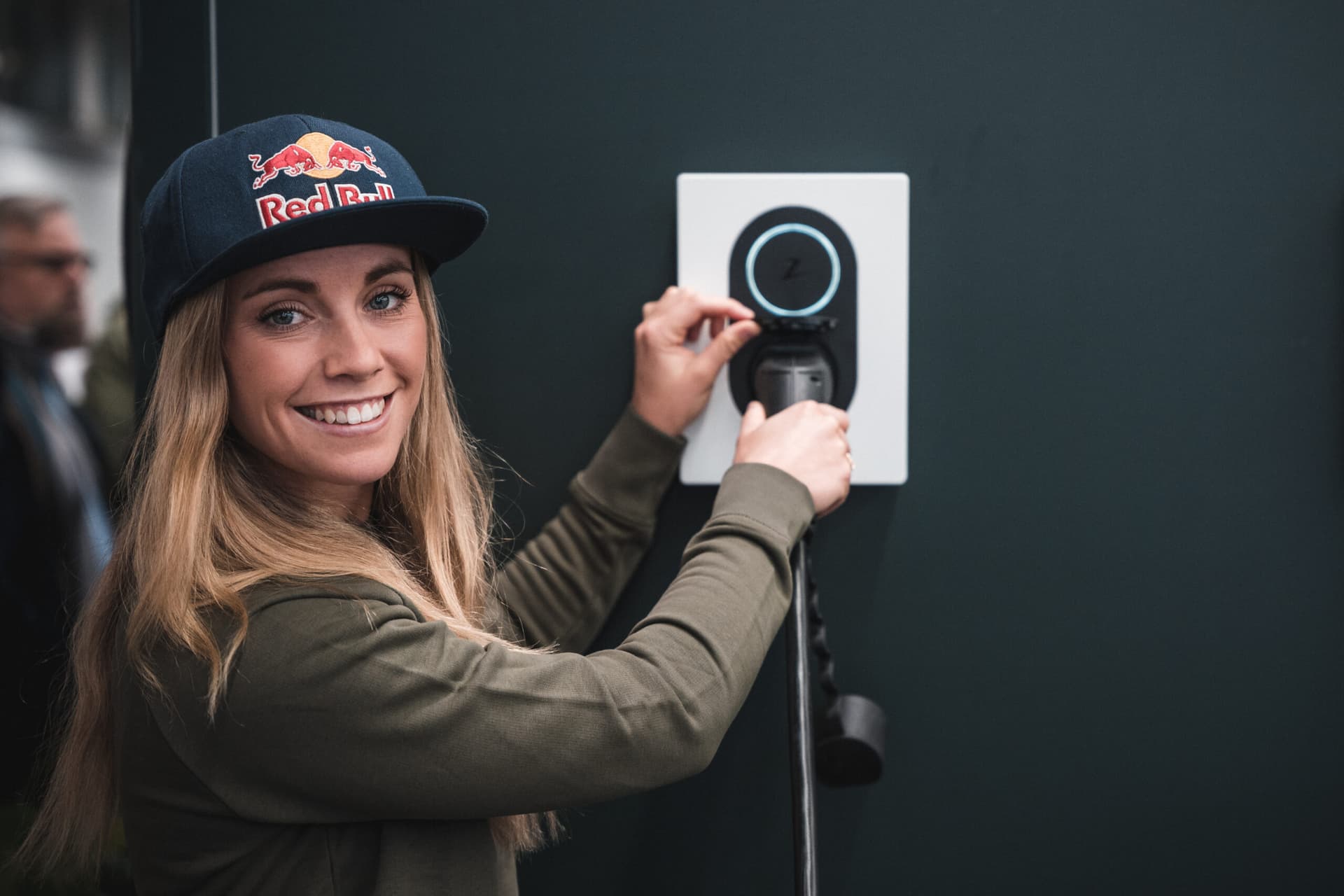 En kvinna med keps ler mot kameran samtidigt som hon kopplar in en laddkabel i en Zaptec Go-laddbox som är monterad på en vägg.