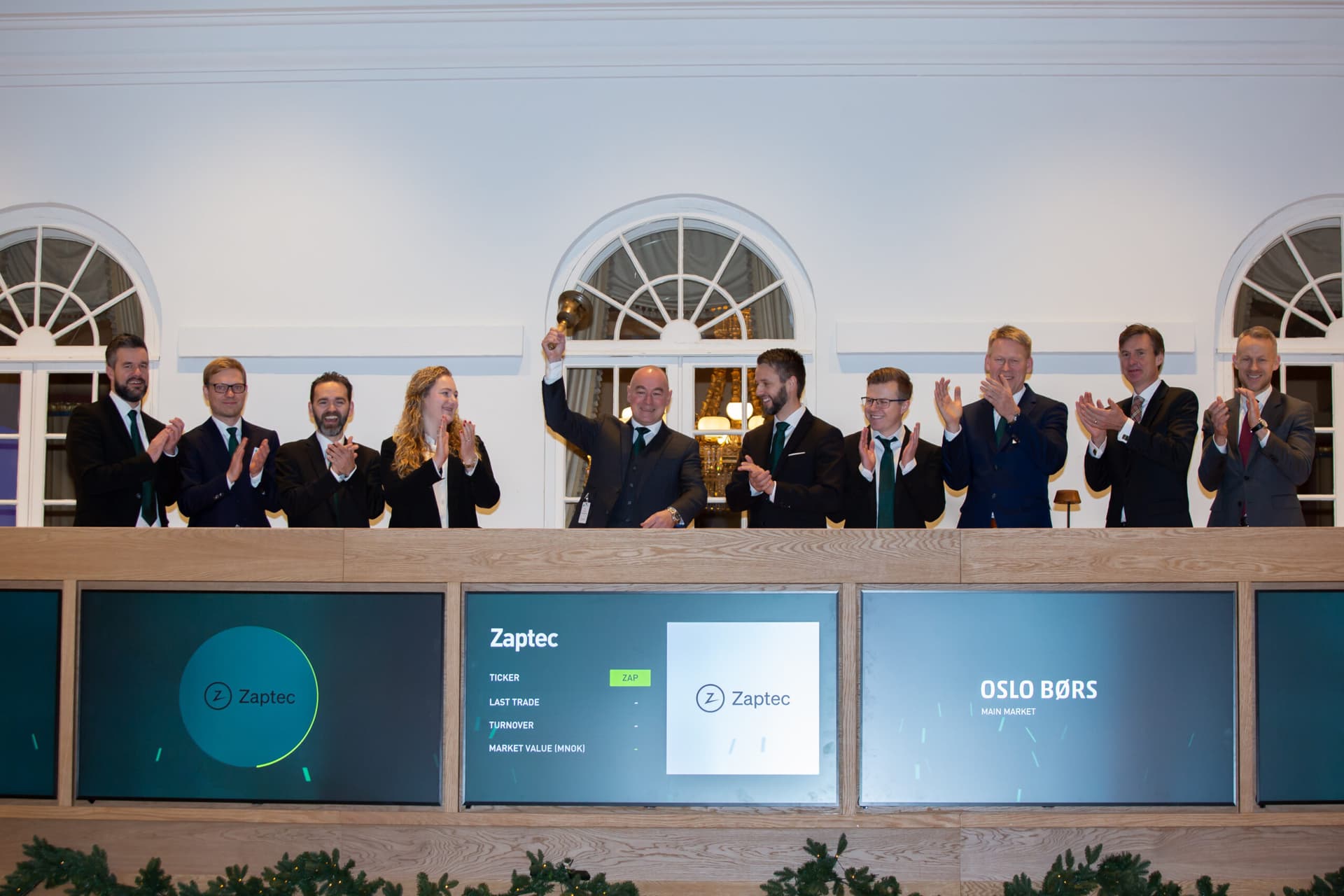 Het team van Zaptec viert de beursnotering op de Oslo Børs. Een man in het midden luidt de bel terwijl zijn collega's in pakken en met groene stropdassen enthousiast applaudisseren.