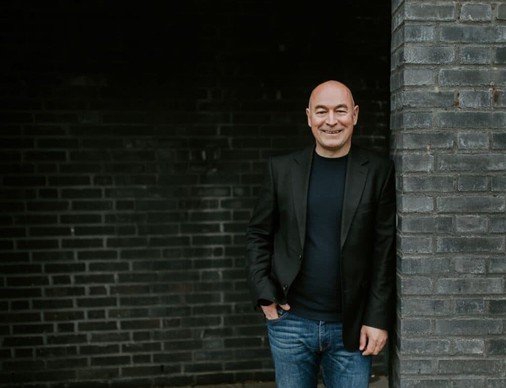 En mann i svart blazer og jeans står smilende mot en mørk teglesteinsvegg med den ene hånden i lommen.