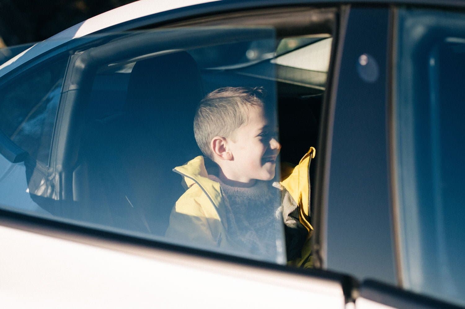 Et barn i gul jakke sitter i baksetet på en bil og ser ut av vinduet med et smil. Sollys lyser opp ansiktet, og refleksjoner vises i bilruten.