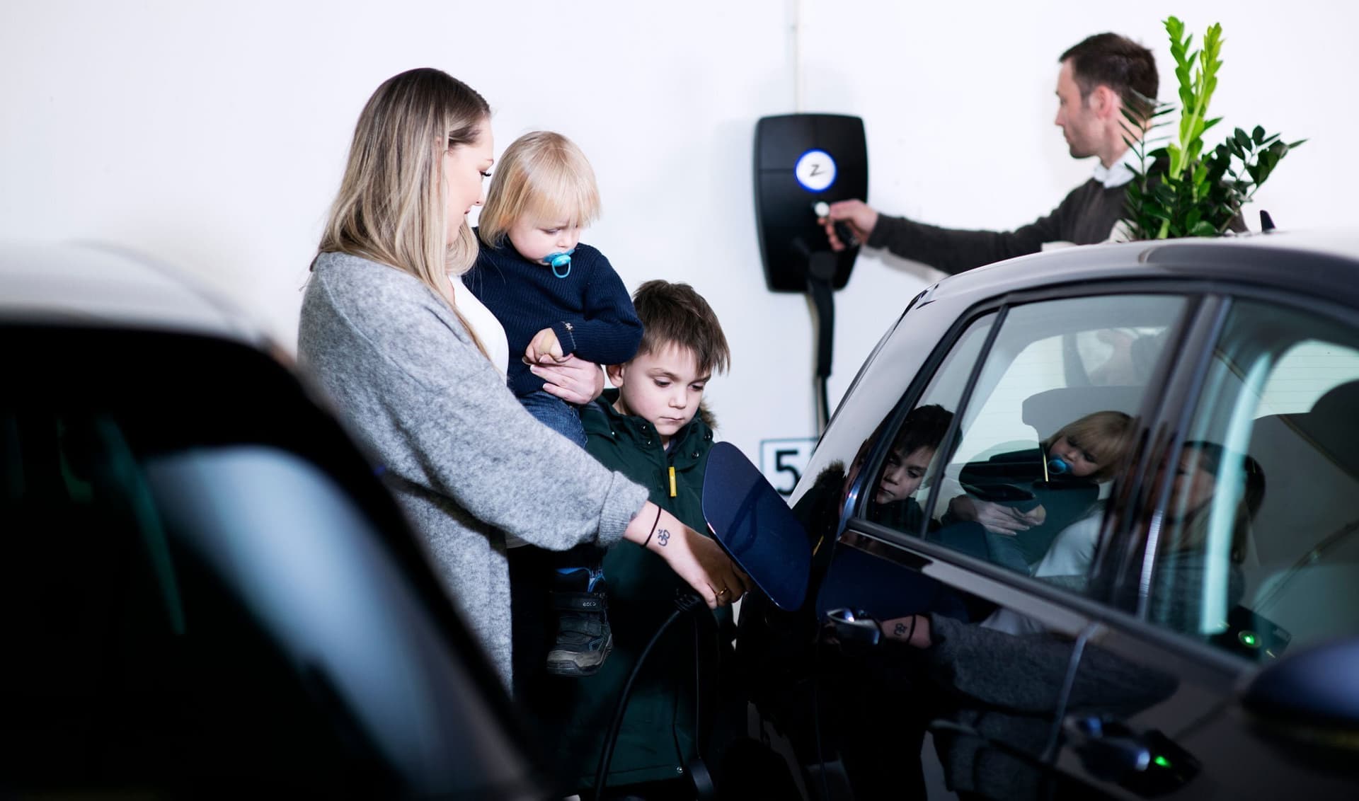 en kvinna i grå tröja med ett barn på armen i mörka kläder och blå napp står tillsammans med en mörkhårig pojke och kopplar in en laddkabel i en svart elbil, bakom elbilen sitter en Zaptec Pro laddare som en man i mörk pullover håller upp en RFID bricka mot.