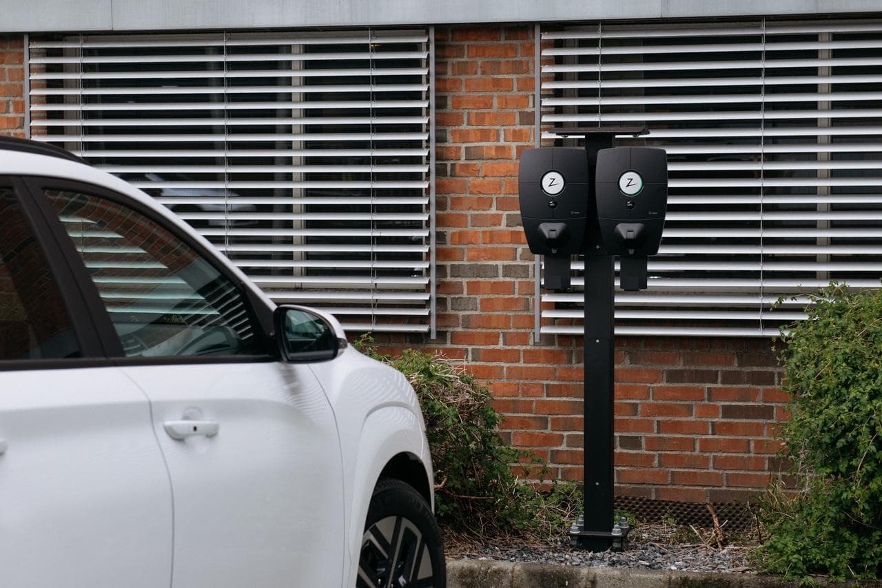 En vit bil står parkerad på en parkering vid en tegelfasad med Zaptec Pro laddare monterade på stolpe