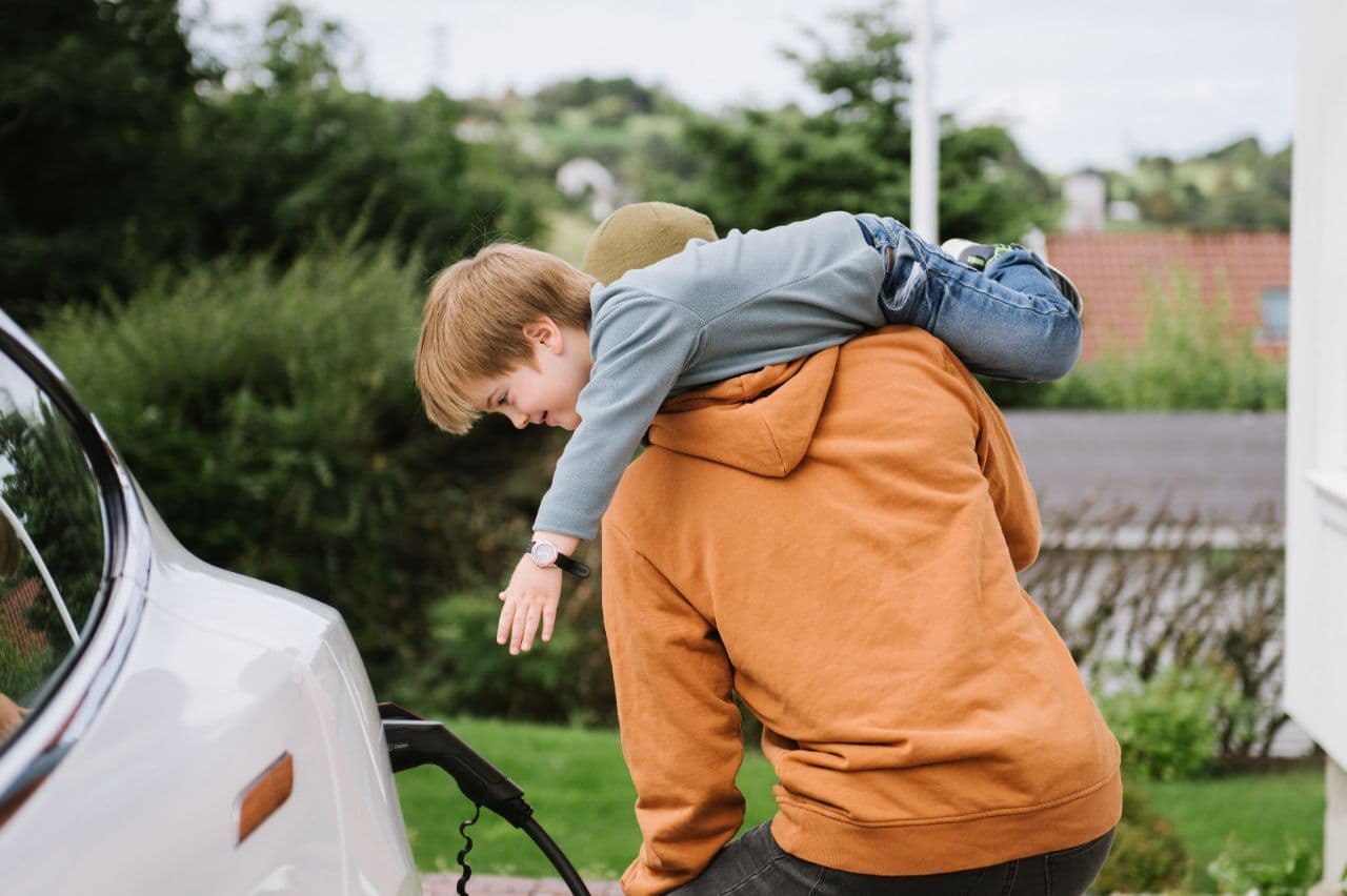 En person i oransje hettegenser lader en hvit elbil mens et barn ligger lekent over den voksnes skuldre og strekker seg mot ladekabelen.