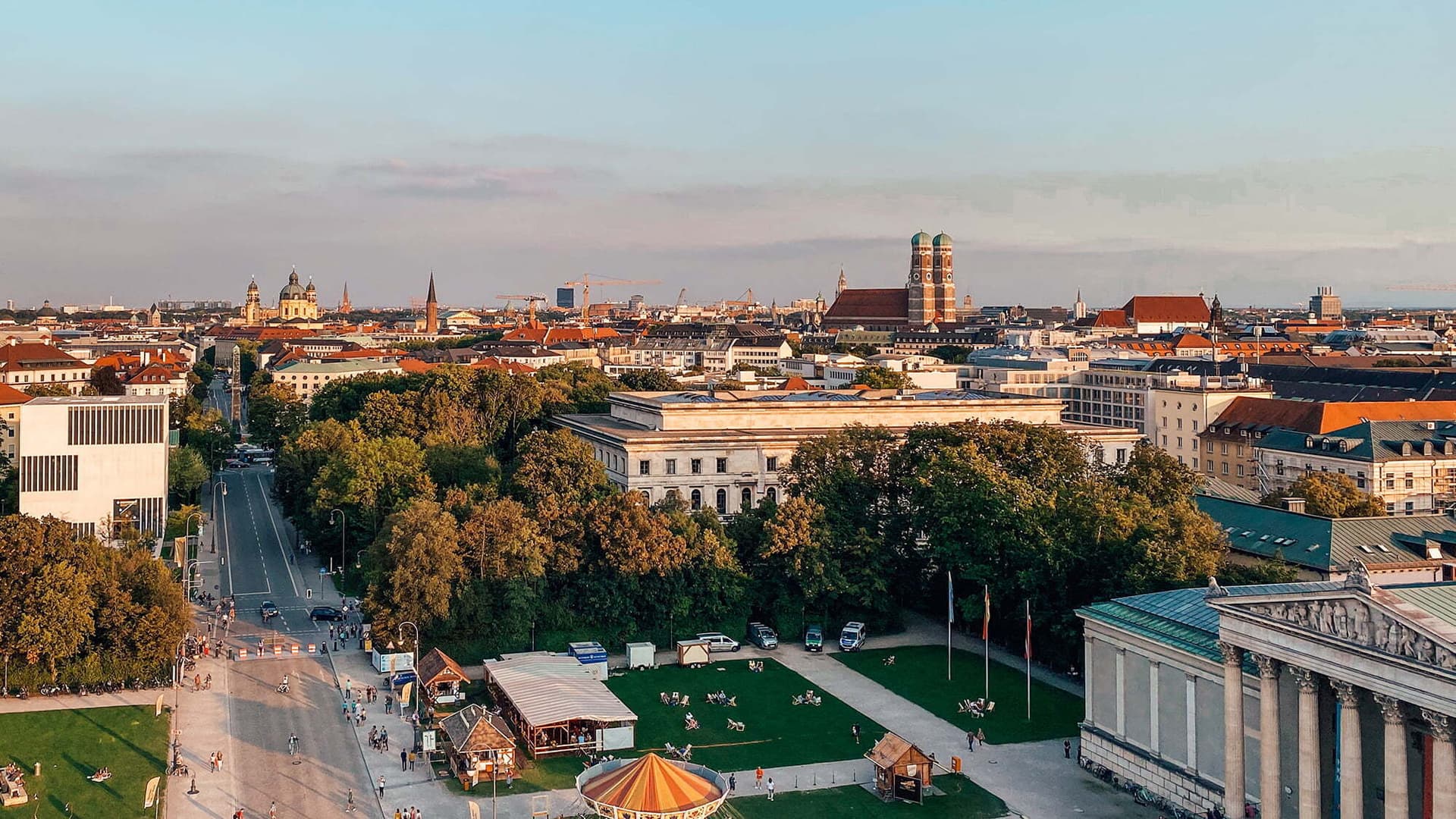 Dronesikt over München med grønne parker, historiske bygninger og byens karakteristiske tvillingtårn i Frauenkirche i bakgrunnen. Folk nyter ettermiddagssolen i parkområdet.