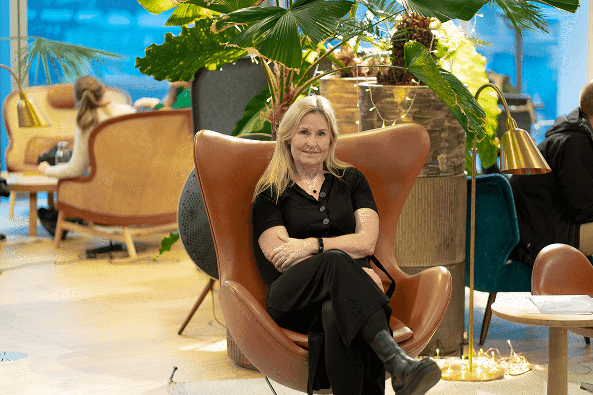 En kvinne med blondt hår og svart antrekk sitter og smiler i en moderne lenestol omgitt av grønne planter og stilige møbler i et lyst loungeområde.