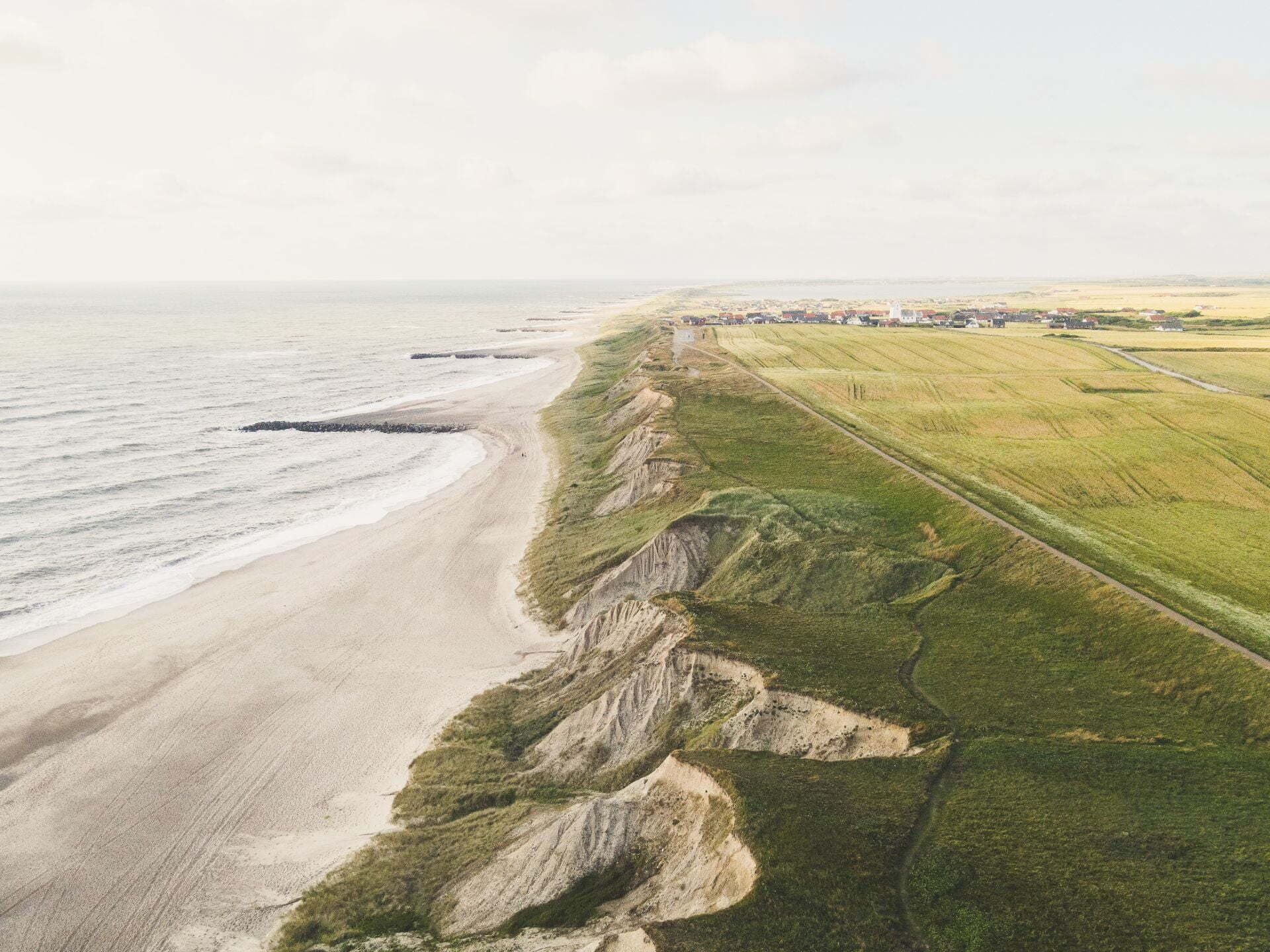 Luftfoto av en kupert kystlinje der sandklipper møter havet, med grønne jorder som strekker seg innover landet under en lett skyet himmel.