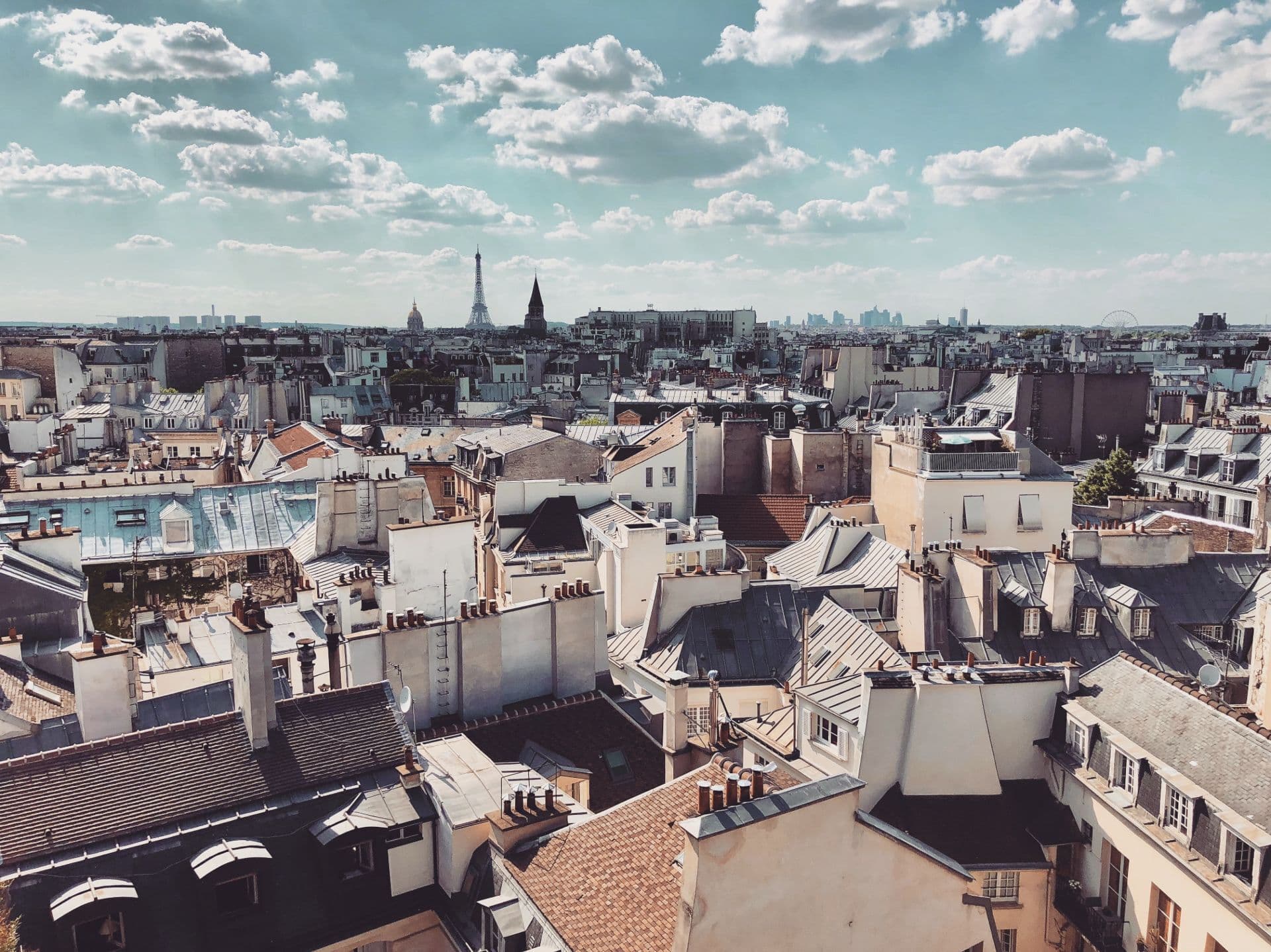 Een panoramisch uitzicht over de daken van Parijs, met zijn typische schoorstenen en zinken daken. In de verte rijst de Eiffeltoren op tegen een blauwe lucht met zachte wolken, terwijl de skyline van La Défense zichtbaar is aan de horizon. De charme van de Parijse architectuur komt tot leven in het warme zonlicht.