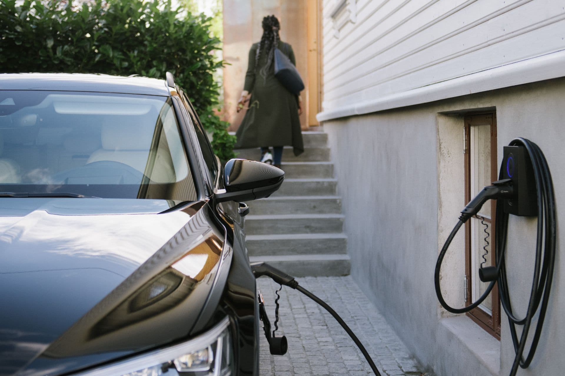 En svart elbil lades med en Zaptec Go-lader som er montert på den hvite trefasaden til et hus. En person i grønn frakk går opp trappen mot inngangsdøren med en svart veske.