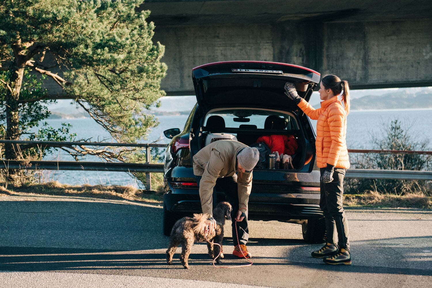 Et par står ved bakluken på en svart bil parkert ved vannet. Mannen med grå lue bøyer seg ned for å feste båndet på en brun hund, mens kvinnen i oransje jakke holder opp bagasjeluken.
