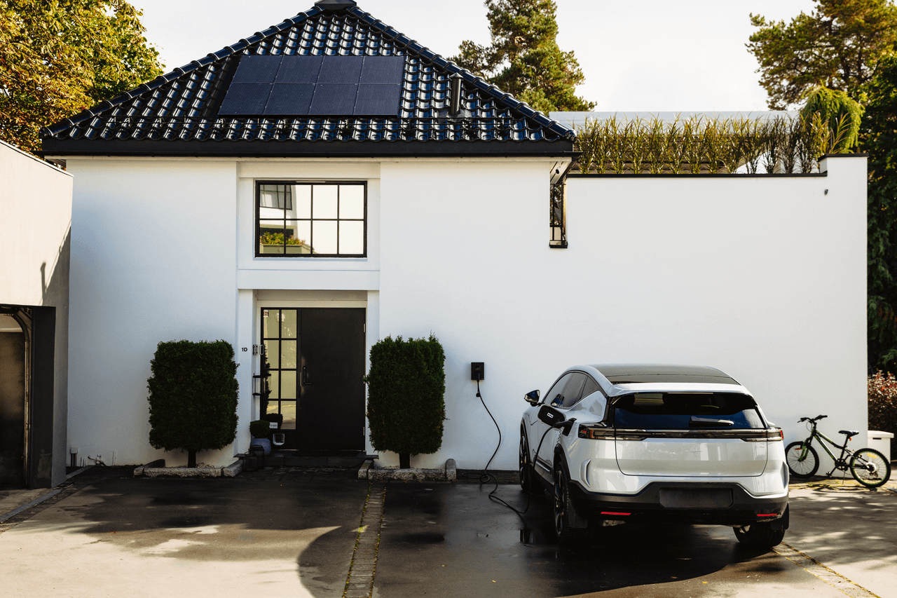 Weißes Haus mit schwarzem Dach, auf dem eine Solaranlage ist. Vor dem Haus lädt ein E-Auto an einer Wallbox