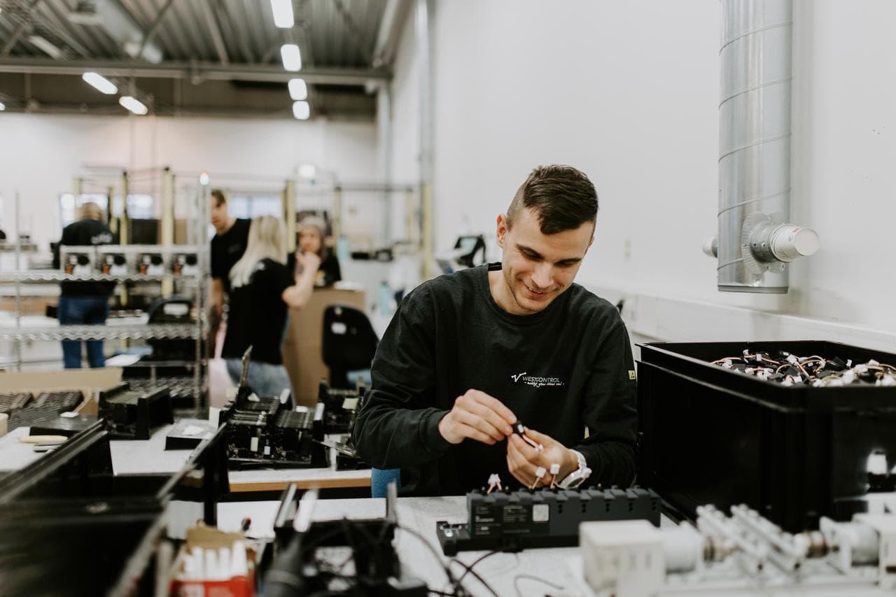 En smilende mann jobber med elektronikkmontering ved en arbeidsstasjon i et lyst og moderne industrilokale. I bakgrunnen sees flere kolleger og komponenter.