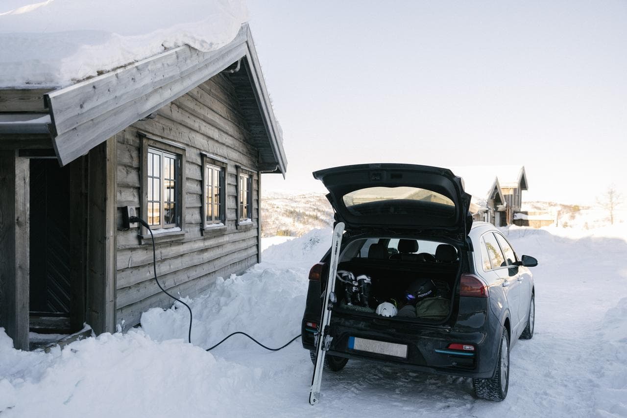 En svart elbil lades med en Zaptec Go-lader montert på veggen til en snødekt tømmerhytte. Bakluken er åpen og et par ski står lent mot bilen i det vinterlige fjellandskapet.