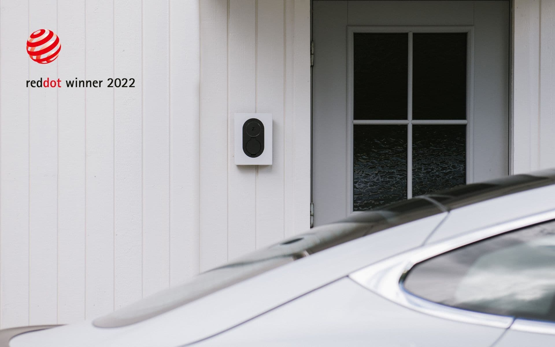 Een witte Zaptec Go-laadpaal is gemonteerd op een witte houten muur naast een glazen deur. Een zilveren elektrische auto staat geparkeerd ervoor, waarbij de reflectie van de omgeving zichtbaar is op het dak. In de linkerbovenhoek van de afbeelding staat het logo 'Red Dot Winner 2022'.
