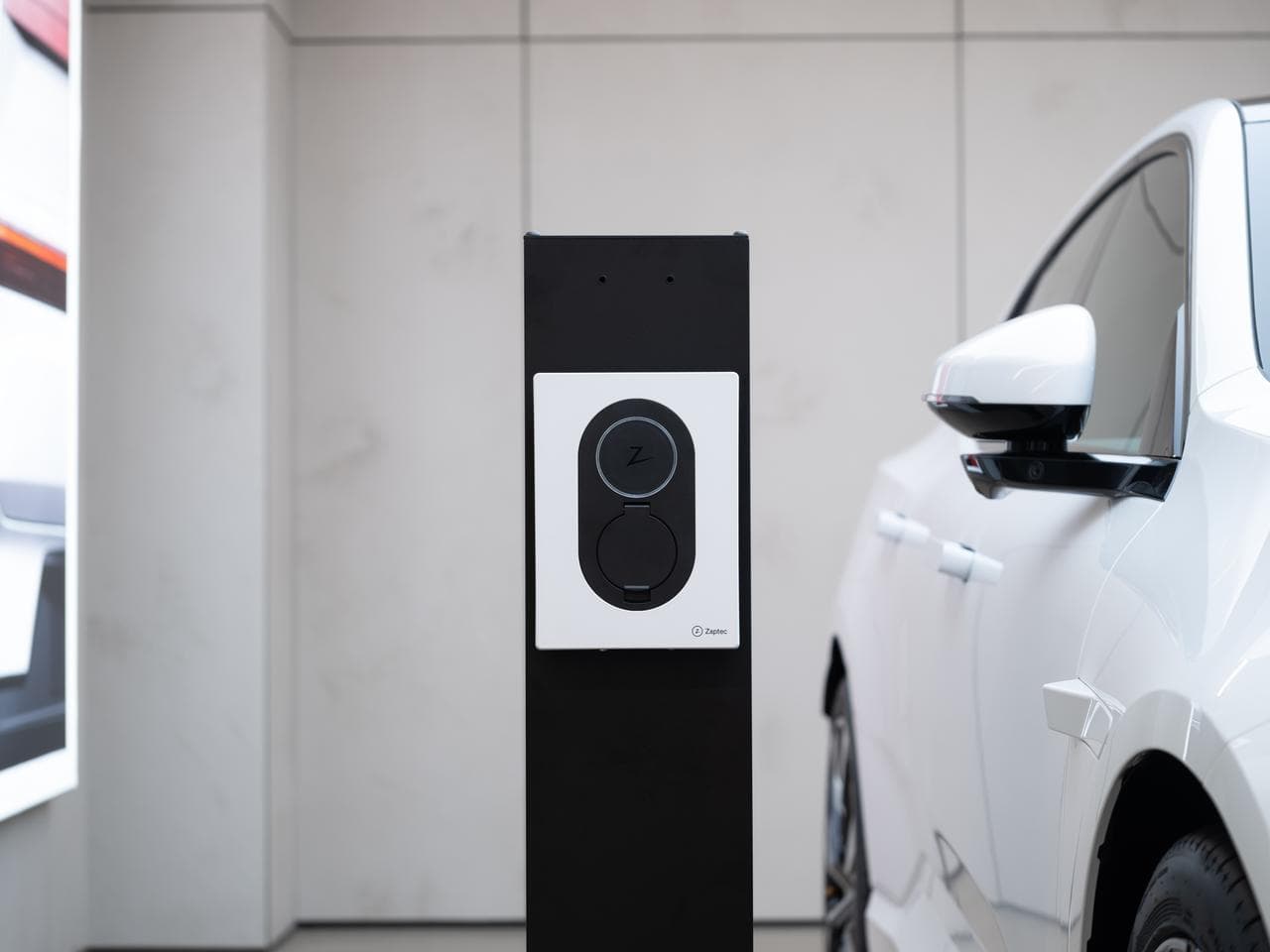 En Cloud White Zaptec Go-lader er montert på en svart søyle i et showroom, ved siden av en hvit elbil.