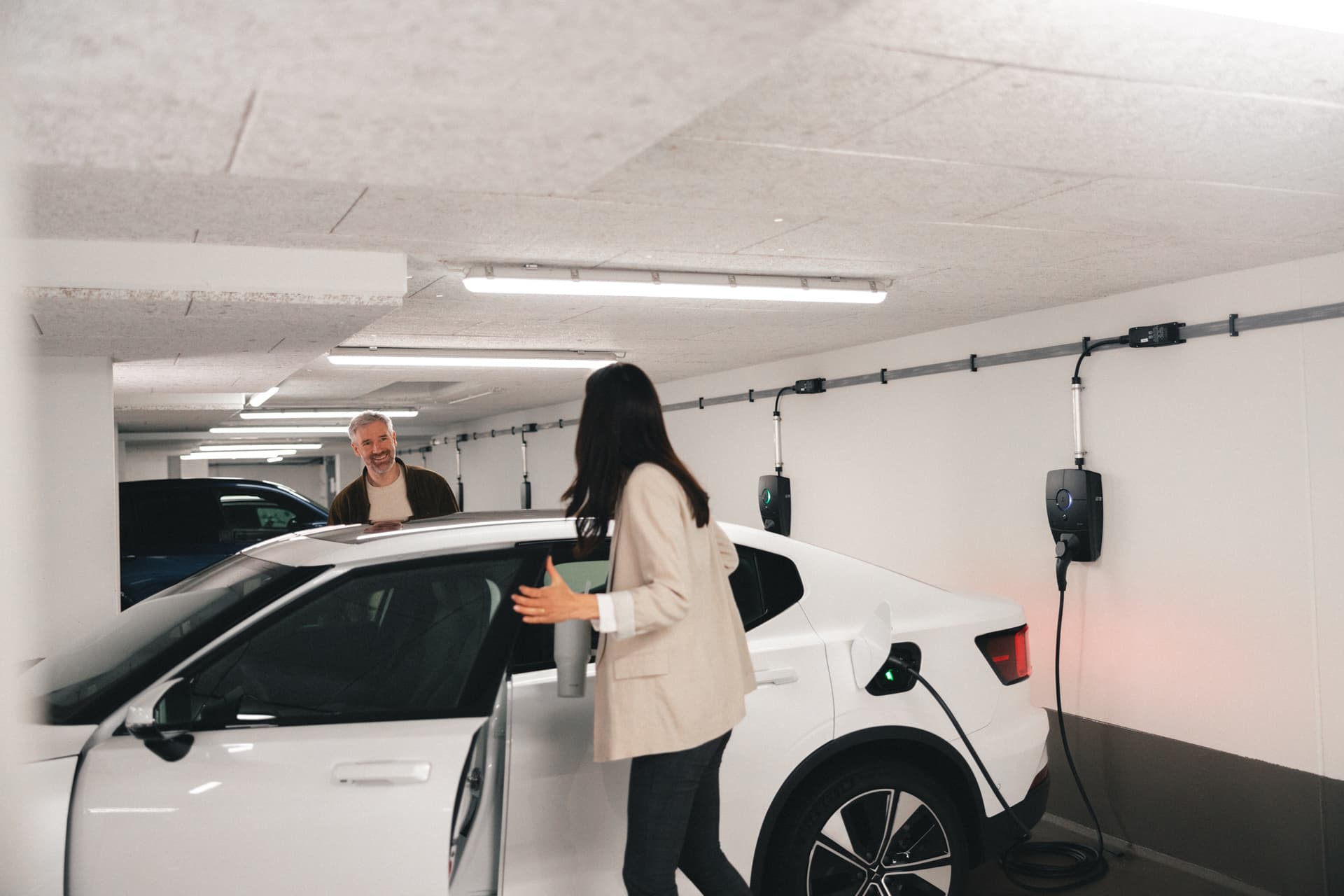 En kvinna står bredvid en vit elbil som laddas med en Zaptec Pro laddare i ett parkeringsgarage. En leende man syns i bakgrunden.