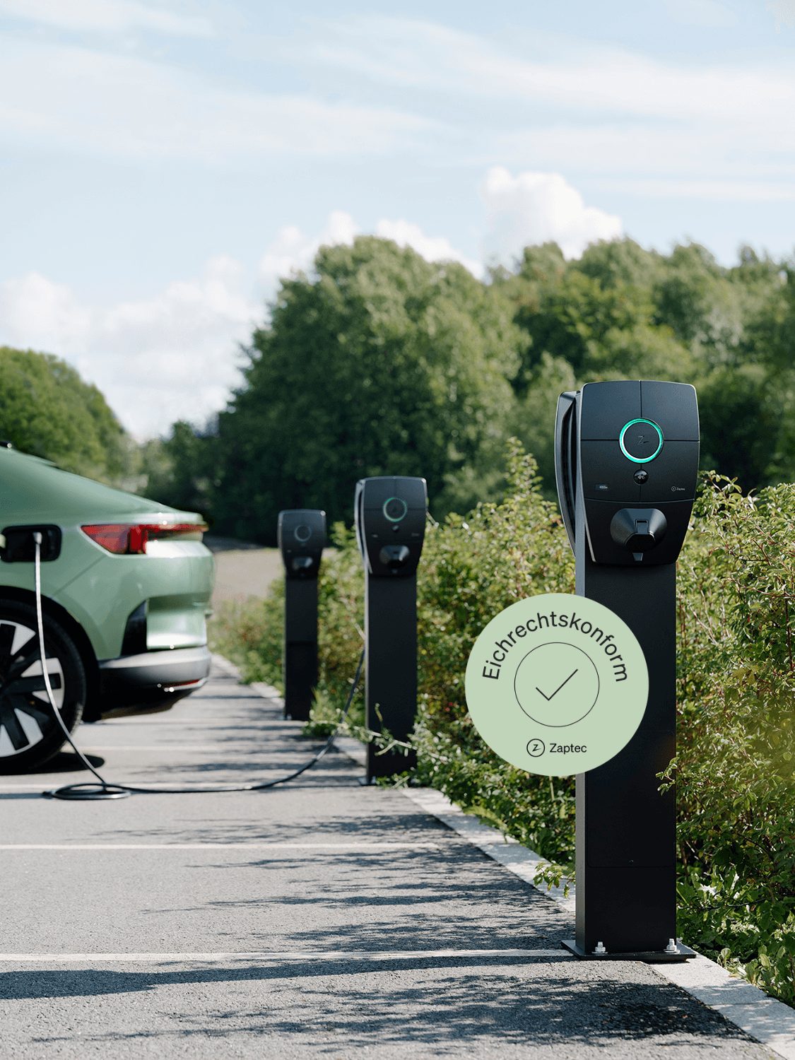 Drei Zaptec Pro Ladestationen auf Säulen im Parkplatz, grünes Auto lädt an erster Station, Grünflächen im Hintergrund.