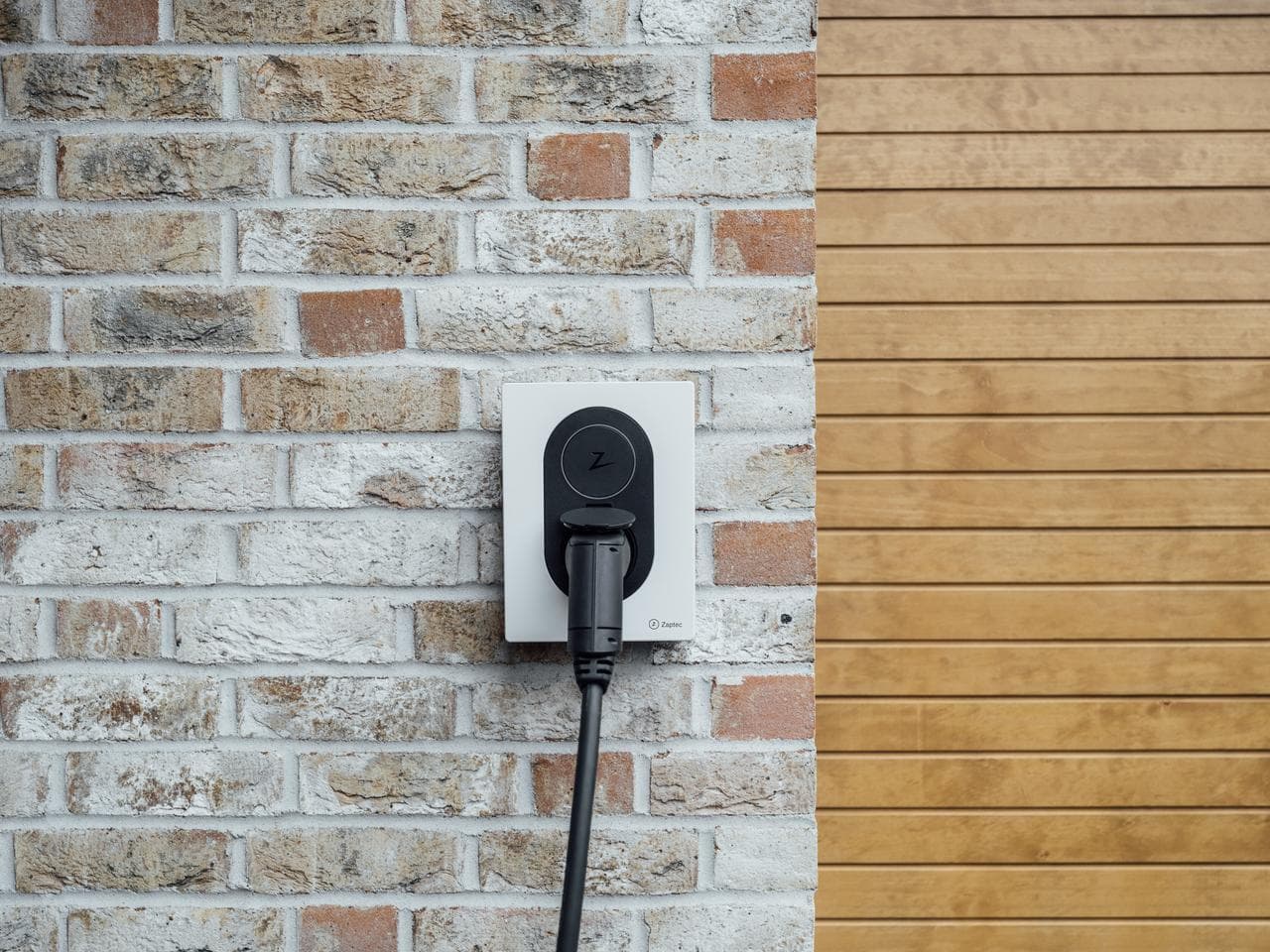 En Cloud White Zaptec Go-lader med tilkoblet kabel er montert på en fasade med lys murstein og trepanel.