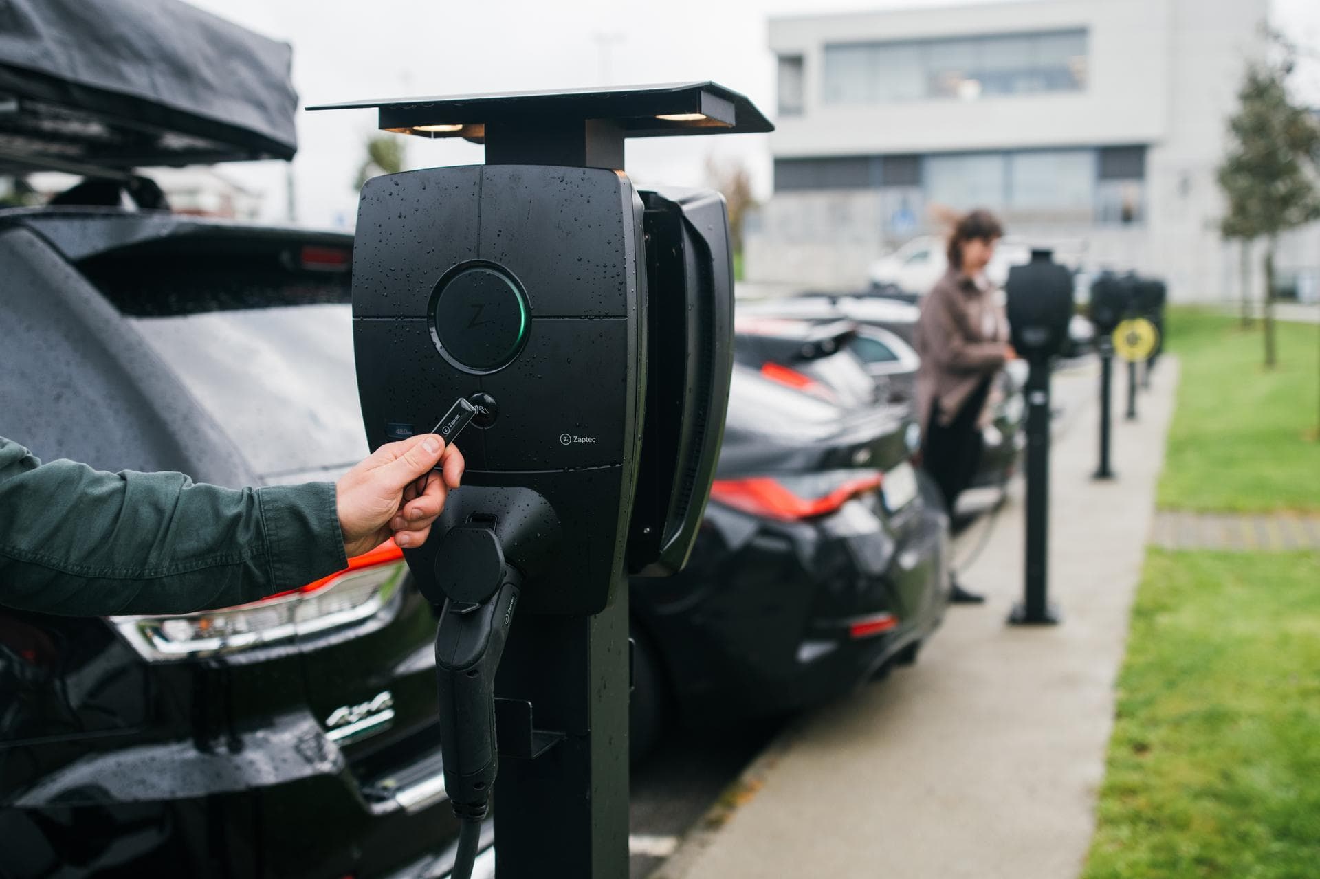 Svara Zaptec Pro MID laddboxar sitter monterade på stolpar längs en parkering där flera bilar står parkerade. En kvinna i brunt kopplar i sin laddkabel längre bort och närmast syns en person som läser av en RFiD bricka mot den närmaste laddboxen.