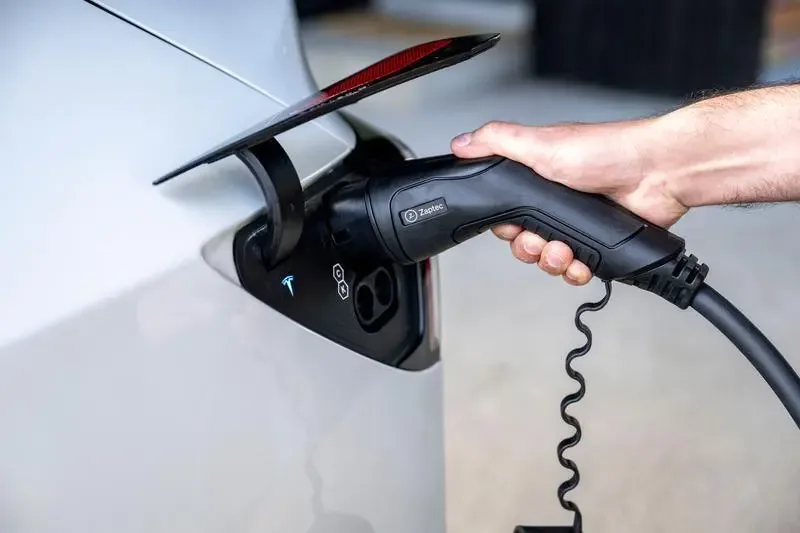 En hånd holder en ladekontakt inntil ladeuttaket på en grå elbil.