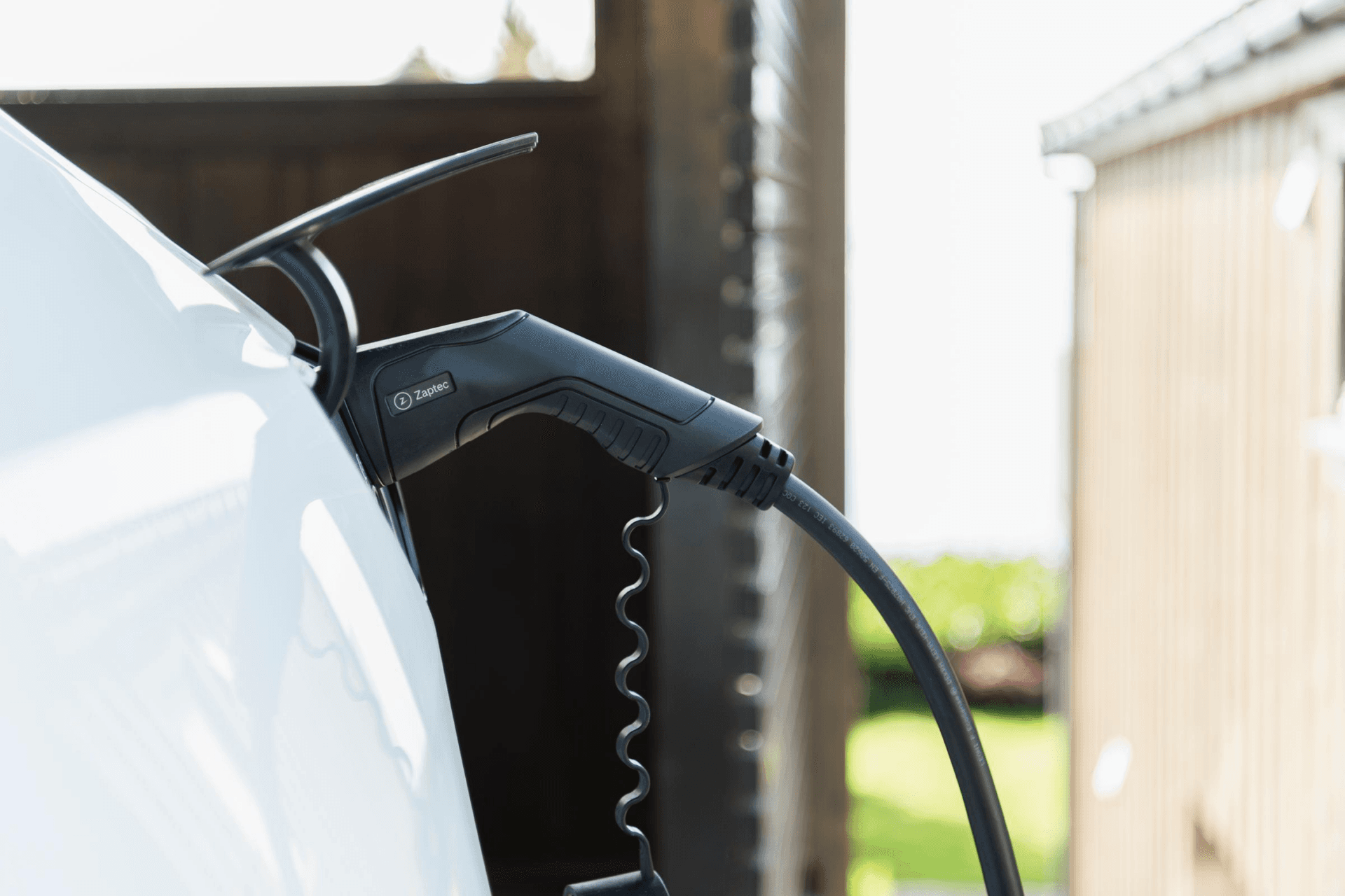 Nærbilde av en ladekabel koblet til en hvit elbil, med sollys som skinner inn mellom en trekonstruksjon og bilen.