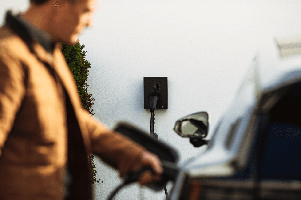 A man in a brown jacket is charging his car with an Asphalt Black Zaptec Go 2 charger mounted on a white wall. The focus is on the charger in the background while the man and the car are slightly out of focus in the foreground.