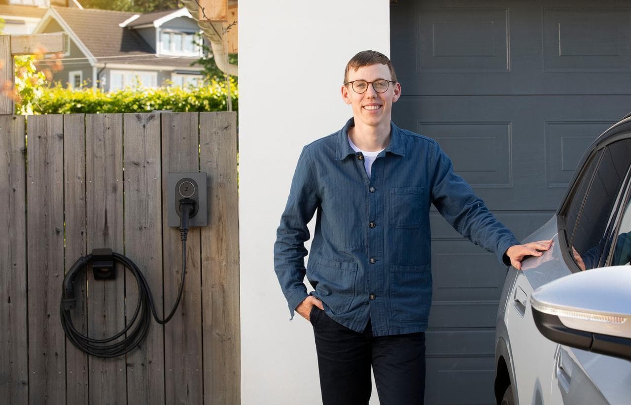 En mann i blå jakke står smilende ved siden av en grå elbil utenfor en garasje. På tregjerdet ved siden av er en Rock Grey Zaptec Go-lader montert med ladekabelen hengt opp ved siden av.